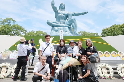 8名の東友会代表が平和祈念像を背景に並んでいる。1名は車いすに座り、花束を抱えている。