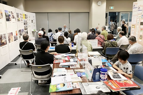 原爆展パネルの掲示されたついたてに挟まれた空間で、並べられた折り畳み椅子に座った20人ほどの参加者が被爆証言を聞いている。手前には書籍などが並べられたテーブルが置かれており、一人が本を広げて読んでいる。