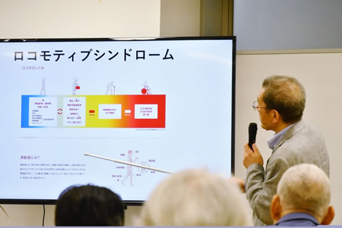 モニタに表示された図を、指示棒も使いながら向山医師が解説をしている。表示されているのは、「ロコモティブシンドローム」の文字の下に図と説明文のあるもの(内容は写真では判読不可)。