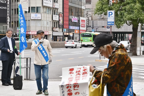 たすきをかけて署名板を手に持った被爆者が、ペンを探しているのか腕に提げたバッグを覗きながら、もう一方の手を使って探っている。その奥で、たすきをかけた被爆者が、マイクを持って道行く人に訴えている。その隣には、一方の手で東友会ののぼりを持った人が立っており、もう一方の手に持ったスマートフォンを見ている。