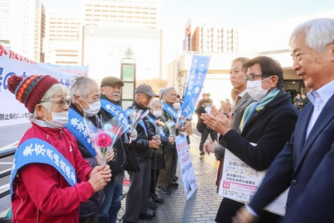 タスキをかけた被爆者が横一列に並び、小さい花束を持っている。花束を贈った人たちがその正面に並んでおり、拍手をしている。