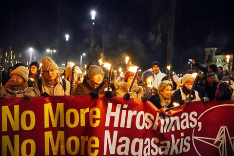 「No More Hiroshimas, No More Nagasakis」と書かれた相談膜を、10数名が横に並んで広げ持っている。片手に松明を持っている人もいる。その後ろに人の列が続いており、いくつもの松明の灯も見える。