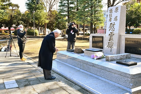 慰霊碑のに向かって、東京都原爆被害者協議会の役員1名が手を合わせ頭を下げている。その様子をテレビカメラがとらえている。マイクのつけられた長い棒を支え持つ撮影スタッフも写っている。