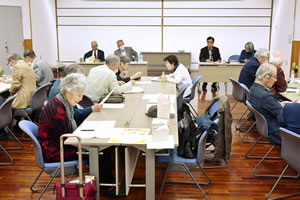 広い部屋、並べられた着席した参加者が、手元の資料を見るなどしながら、前方の机に座って話す人の話を聴いている。
