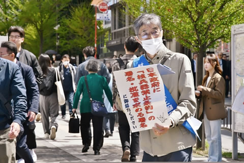タスキを掛けた被爆者が街頭に立ち、署名板から下げた「日本政府に『核兵器禁止条約』の署名・批准を求める署名に御協力ください」と大きく書かれた紙を胸の前に掲げながら、通行人に署名への協力を呼び掛けている。