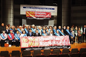 舞台の上に集まった参加者たち。最前列の人たちが、「No More Hiroshimas, No More Nagasakis」「Nihon Hidankyo 2024 Nobel Peace Prize」の英文と折り鶴のイラストか書かれた横断幕を広げ持っている。舞台奥の天井からは「2025 ピースアクション in TOKYO」など大きく横書きれた看板がさげられており、看板下部に舞台上の人たちが広げ持っているものと同じ横断幕が付けられている。東友会の参加者は、たすきをかけている。