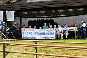 第五福竜丸のエンジンが展示されている屋根の下、タスキを掛けた東友会の参加者が横並びに椅子に座り「核兵器のない平和な世界の実現を」と大きく書かれた横断幕を広げている。うち一人が立って、マイクを使い話している。。