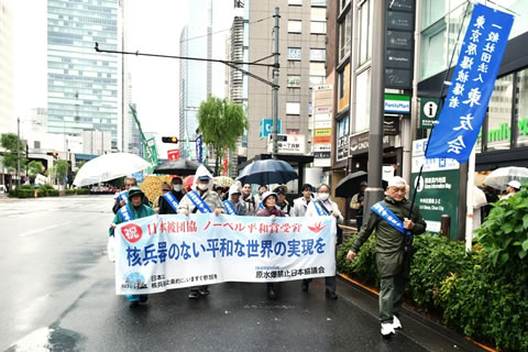 タスキを掛け、東友会ののぼりを掲げた人の後ろに、「祝 日本被団協ノーベル平和賞授賞 核兵器のない平和な世界の実現を」と書かれた横断幕を広げ持つ5人を先頭にした行進が続いている。参加者は傘を差したり雨合羽を着たりしている。