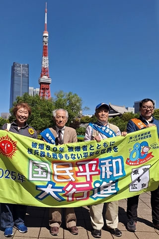 「国民平和大行進」など書かれた横断幕を、タスキを掛けた2人の被爆者を真ん中にして4人が広げ持っている。端のひとりは「通し行進者」のタスキを掛けている。背景には、晴れた空にそびえる東京他w。