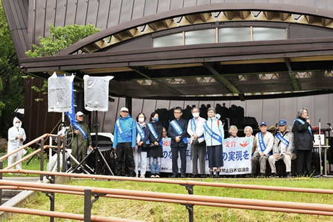 第五福竜丸のエンジンが展示されている屋根の下、東友会のタスキを掛けた被爆二世たちが横並びに立っている。その後ろに東友会のタスキを掛けた被爆者たちが横並びの椅子に座って横断幕を広げている。