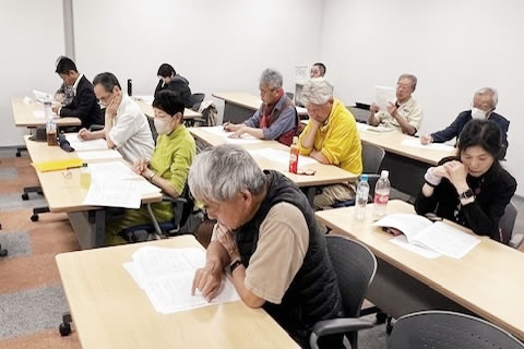 室内、並べられた机に着席した参加者たちが、手元の資料に目を落としている。