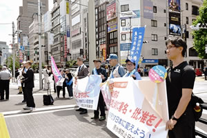 タスキを掛けた被爆者ほか参加者が、通行人に向かって横断幕を広げている。(横からの撮影なので横断幕の文字はよく見えない。)東友会ののぼり、署名を呼び掛けるのぼりも立てられている。