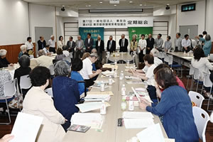 会場前方、「第77回 一般社団法人東友会 第69回 東京都原爆被害者協議会 定期総会」と大きく横書きされた看板が天井近くに下げられている。その下には東友会の旗が下げられている。その手前に、選出された役員が一列の横並びで立っている。他の出席者は、並べられた長机に着席し、役員たちのほうを見ていたり、資料に目を落とすなどしている。