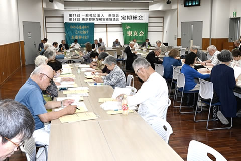 会場前方、「第77回 一般社団法人東友会 第69回 東京都原爆被害者協議会 定期総会」と大きく横書きされた看板が天井近くに下げられ、その下には東友会の旗が下げられている。その手前に会場に向けて並べられた机に、議長などが着席している。他の出席者は机に着席し資料に目を落とすなどしている。