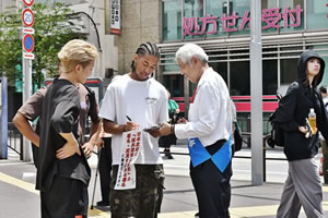 タスキを掛けた被爆者のもつ署名板で、ドレッドヘアを後ろで束ねたTシャツに軍隊風のカモフラージュ柄ズボンを