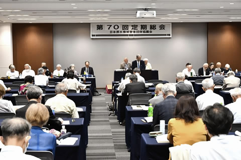 会場前方、天井から「第70回定期総会」と大きく横書きされた看板がさげられている。演台に2人経った1人がマイクを使って話している。他の参加者たちが、並べられた机に着席し、手元の資料に目を落とすなどしながら聴いている。