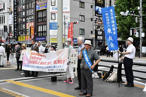 広い歩道で、「日本政府は核兵器禁止条約に署名・批准を」と書かれた横断幕を、歩行者に向けて数人が広げ持っている。そのわきに、意見広告と思われるパネルを持って立っている人がいる。その隣で、タスキを掛けた被爆者がマイクを使って呼びかけている。東友会ののぼりを持っている人も写っている。