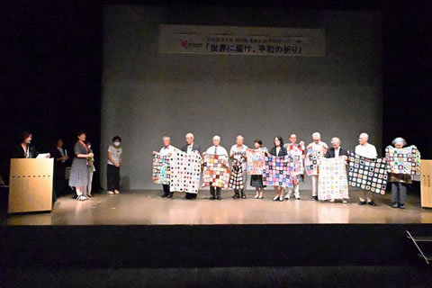 舞台の上に、手編みのひざ掛けを客席に向け広げ持って並ぶ被爆者たち。