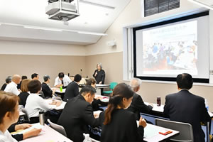 会場前方、天井から「第70回定期総会」と大きく横書きされた看板がさげられている。演台に2人経った1人がマイクを使って話している。他の参加者たちが、並べられた机に着席し、手元の資料に目を落とすなどしながら聴いている。