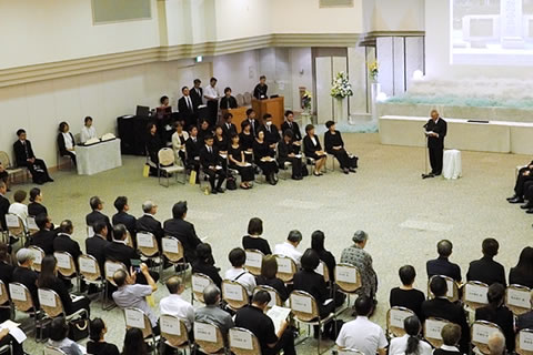 会場前方に天井から下げられた大きいスクリーンに「慰霊碑」の写真が投影されている。スクリーンの手前に、マイクの前に立って手元の紙を読み上げている人がいる。参列者たちが並べられた椅子に座ってそれを聞いている。