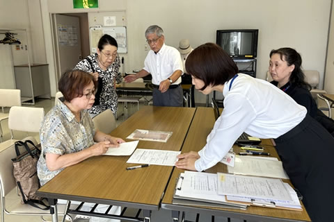向かい合わせの長机に、広島市職員2名と東友会代表1名が向き合っている。うち職員1名は立って身を乗り出すように書類に手を添えており、説明しているように見える。他に東友会代表2名が脇に立って書類を見ている。