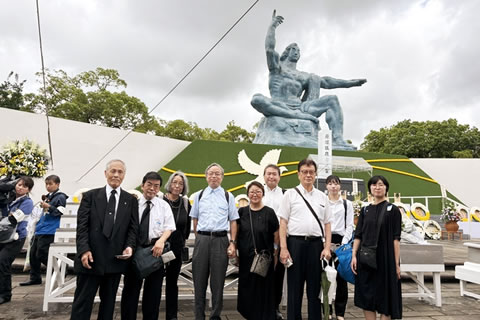 9名の東友会代表が平和祈念像前に置設置された献花台の前に並んで立っている。