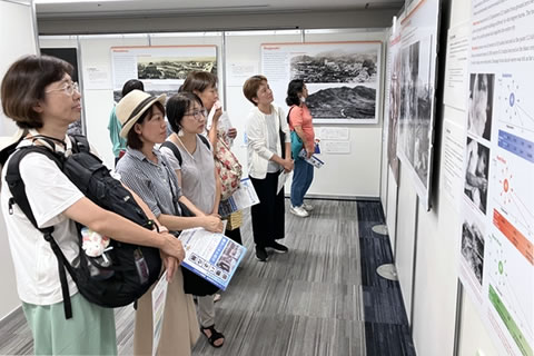 壁に横並びに展示されたパネルをそれぞれに見ている人6人が写っている。