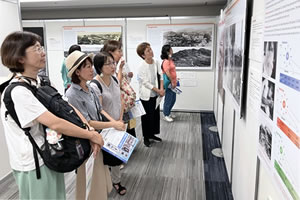壁に横並びに展示されたパネルをそれぞれに見ている人6人が写っている。