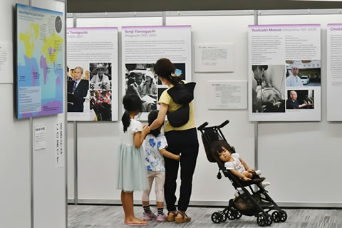 1枚のパ根雨の前に立つ人が、パネルを指さしながら、小学生くらいに見える子どもたち2人と一緒に見ている。パネルの意味はまだわからないであろう小さい子が1人、ベビーカーに乗せられている。