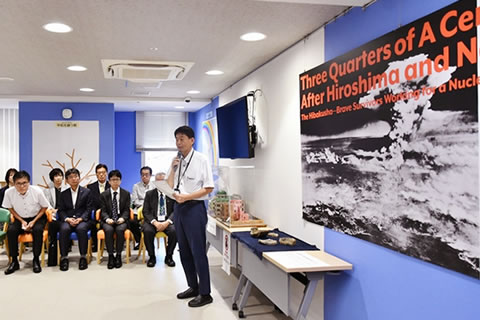 東京都の二宮課長が壁際の展示物の前に立ち、囲むように置かれた椅子に着席する参加者に向かってマイクを使って話している。壁に展示されたパネルも写っている。