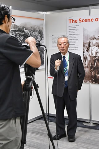 会場内、展示されたパネルを背景にテレビカメラに向かって立ち、マイクを使って話す家島代表理事。