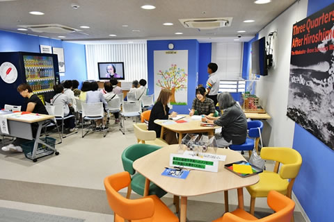 座って話ができる、角の丸い5角形のテーブルと椅子の組が2つある。1つには3人の人が座って話している。液晶テレビの前に椅子が並べられた場所では、10人くらいの人が映像を見ている。