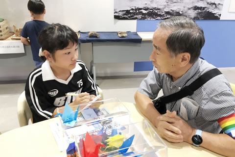 テーブルの席について会話している被爆者1人と小学生に見える子ども1人。子どもはメモ帳とシャープペンシルを持ってメモしている。テーブルの上に折り鶴の入ったアクリルのケースがある。