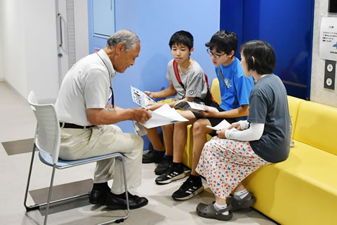 小学生に見える子ども3人がソファに座り、向かいの椅子に被爆者が1人座って話をしている。4人とも手には紙の資料らしきものを持っているが、子どもたちは被爆者が持って示している髪をのぞき込んでいる。