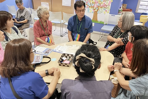 1つのテーブルに8人が座って会話している。目印を首から提げた主催側が3名で、うち2名が被爆者に見える。