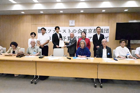 「都民ファーストの会東京都議団 予算要望ヒアリング」と大きく横書きされた紙が壁に張られている。その手前に議員や被爆者が横並びに数人立っており、中央では議員の1人と被爆者の1人が1枚の要望書を両側から持って掲げている。その手前に横並びに置かれた机の席に着席している人たちもいる。