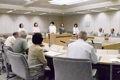 半円形のような形に並べられた机に、東友会の参加者たちが着席している。向かい合う形で横並びに議員らが立っており、うち中央の1人が話をしている。