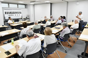 並べられた長机に着席した参加者たちのうち1人が立ち、マイクを使って質問をしている。「地区相談員研修交流会」と大きく横書きされた紙が、会場前方のホワイトボードに張られている。その前の机に講師が座っており、質問に答えようとしている。