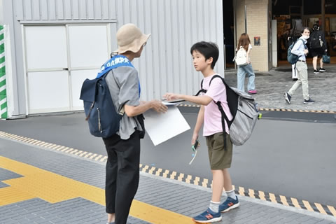 タスキを掛けた被爆者が署名板を持って立っている。子どもが1人、その署名板にペンを持った手を置きながら、被爆者と会話している。