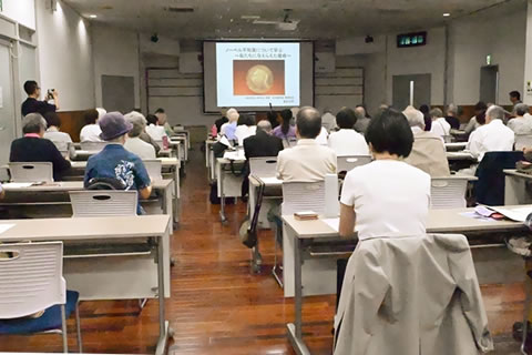 会場前方、天井から下げられた大きいスクリーンに図と説明が投影されている。（写真では文字が小さく判読不可）並べられた長机に着席した参加者がそれを見ながら話を聞いている。