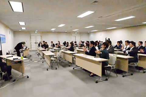 話を終えて頭を下げる証言者に向け、広い部屋に並べられた机に着席した都議たちが拍手をしている。