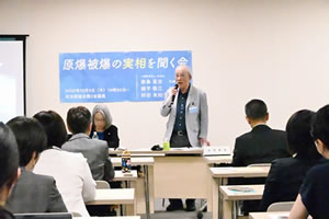 会場の前方で立ち、マイクを使って話をする綿平敬三さん。その背後の壁に、「原爆被爆の実相を聞く会」と大きく横書きされた紙が貼られている。