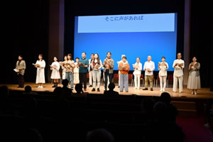 舞台の上に20人ほどの人が朗読台本を胸の前あたりに持ち、客席を向いて立っている。その背後のスクリーンに、「そこに声があれば」の文字が投影されている。