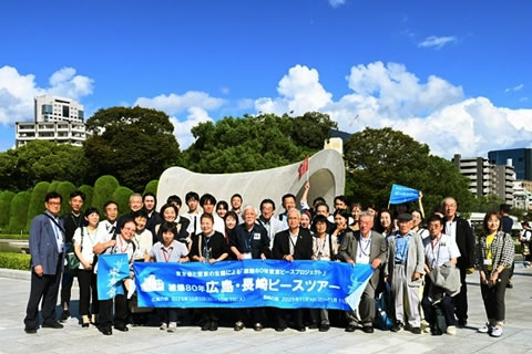 広島平和記念公園の原爆死没者慰霊碑を背景にした参加者の集合写真。「被爆80年 広島・長崎ピースツアー」と書かれた横断幕を広げ持っている。