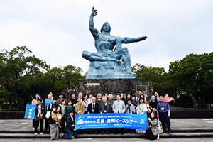 平和祈念像を背景にした参加者の集合写真。「被爆80年 広島・長崎ピースツアー」と書かれた横断幕を広げ持っている。