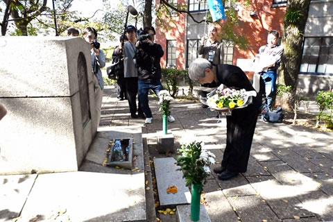 長崎市立山里小学校nいある「あの子らの碑」の前で、花束を持ち腰を折るように頭を下げる人。その様子を、�テレビクルーたちが撮影している。