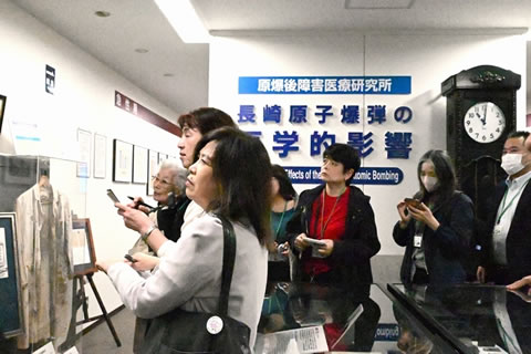 展示された資料を見る参加者たち。見ている展示物自体は写っていないため不明。壁に「原爆後障害医療研究所 長崎原子爆弾の医学的影響」の文字がある。また、長崎原爆がさく裂した時刻である11時2分を指して止まっている柱時計が置かれている。