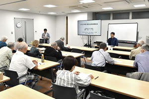 並べられた長机に、参加者が着席している。会場前方の長机に講師が着席している。前方の端のほうで、司会と思われる人がマイクを持って立って話をしている。