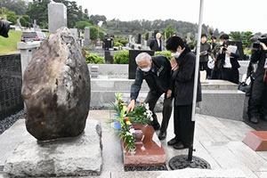 「原爆被害者の墓」の前に立って手を合わせている人と、足元の献花台に花を置いている人がいる。その様子をテレビクルーらが撮影している。