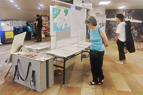 壁には飛行中のB-29爆撃機を上からとらえた航空写真の大型パネルなどが展示されている。室内に立てられたついたてと、それを挟む形で置かれた長机には地図や細かい文字の展示物がある。数名の来場者が展示物を見ている。